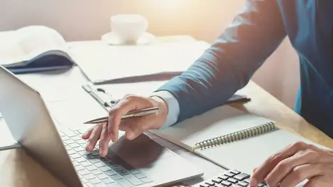 Um homem de terno fazendo cálculos com auxílio de uma calculadora, e no laptop.